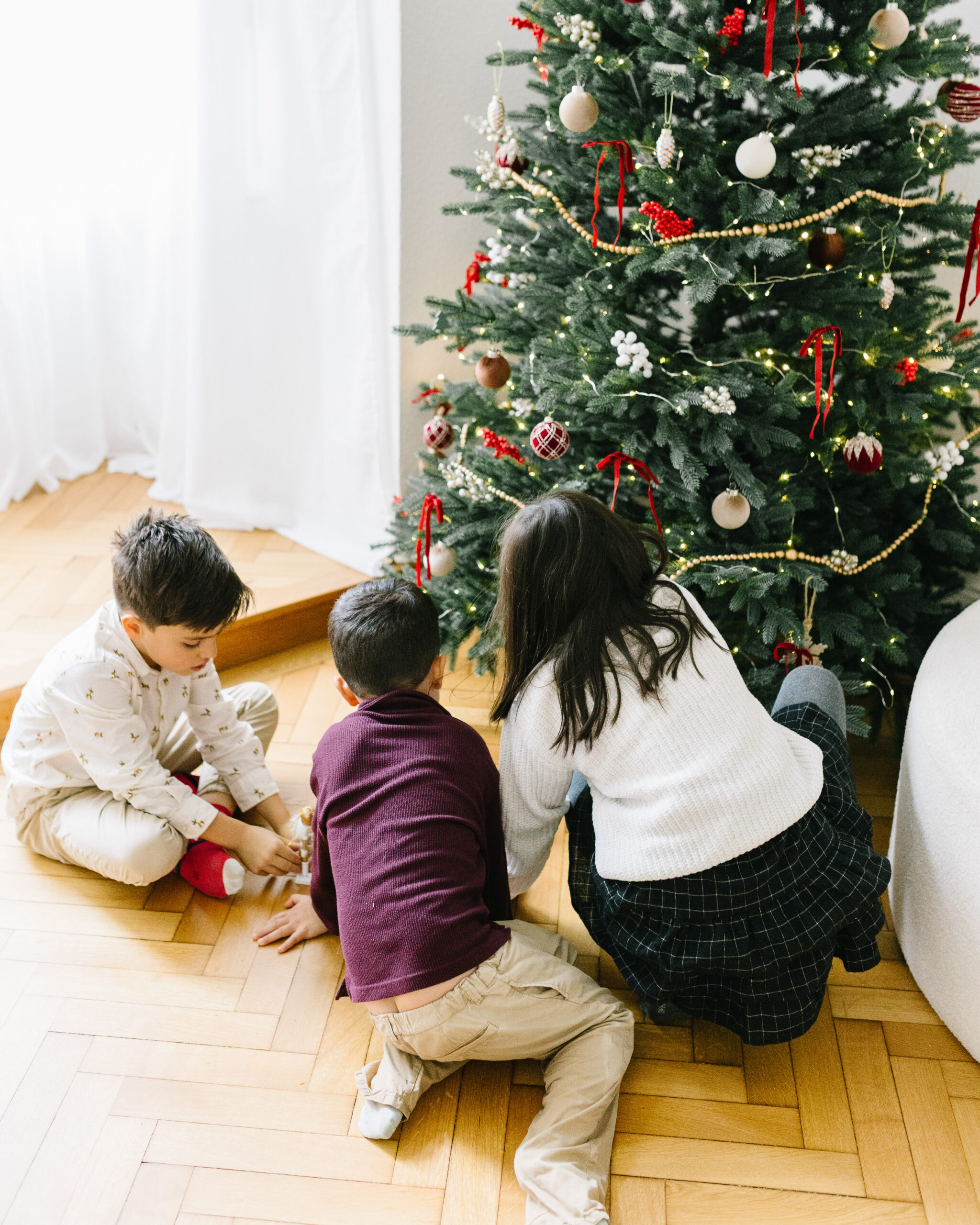 Drei Kinder sitzen gemeinsam auf dem Holzboden vor einem festlich geschmückten Weihnachtsbaum.