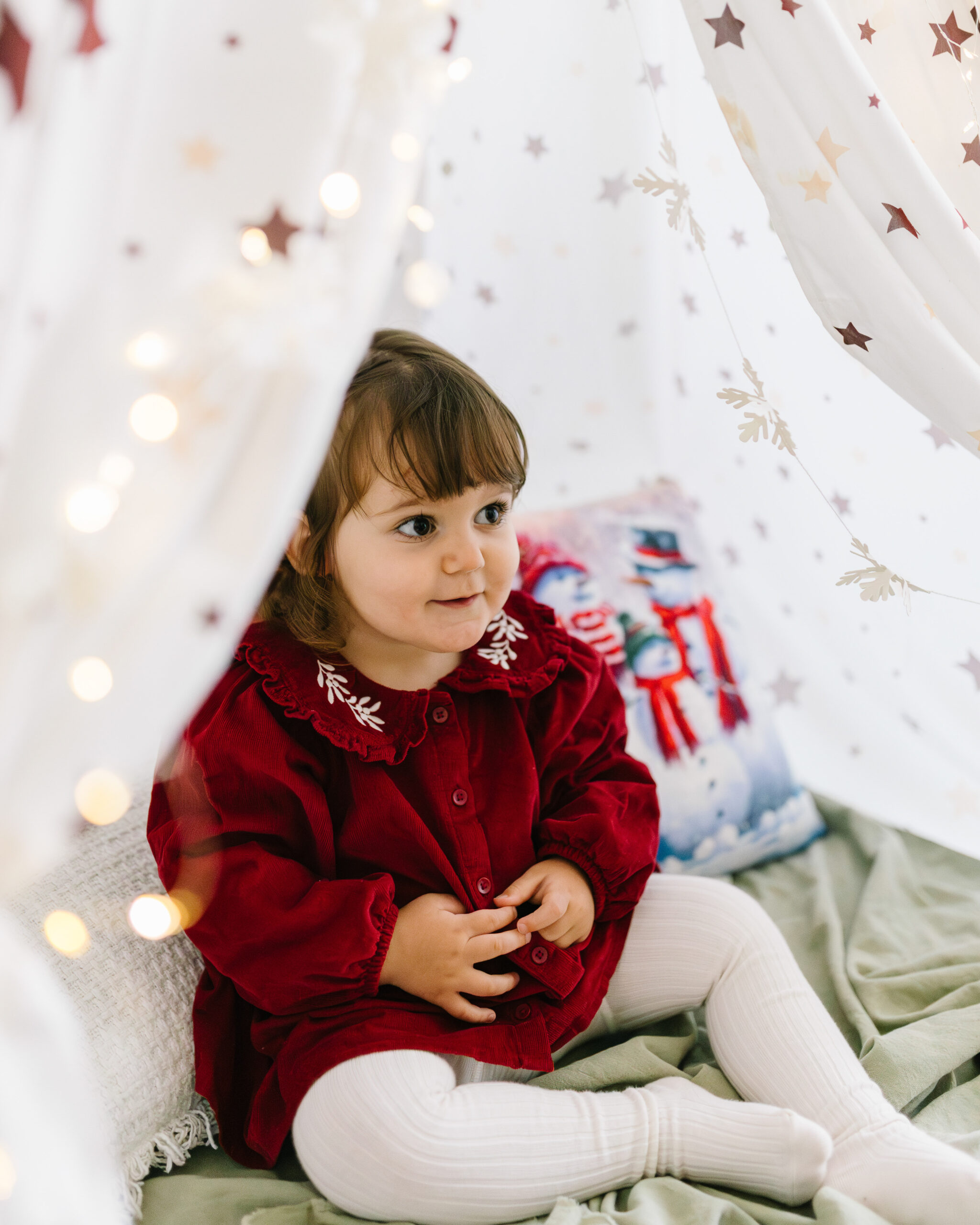 Kleines Mädchen in einem roten Kleid sitzt in einem weihnachtlich dekorierten Spielzelt mit Lichterketten.