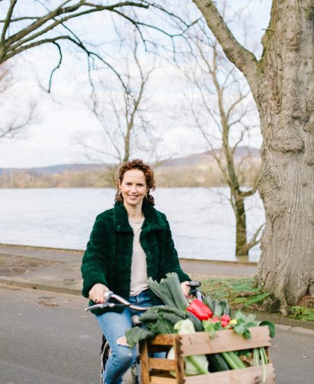 Curly-haired woman riding a bicycle by the Rhein River in Bonn, Germany, smiling at the camera.