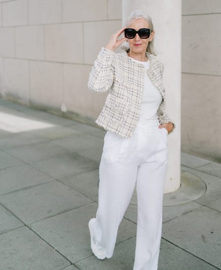 A white haired woman in a white suit walking on the street and holding her sunglasses.